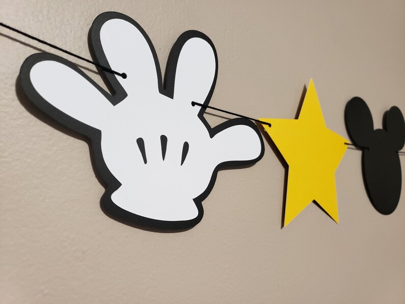 Puede incluir: Una mano de Mickey Mouse en blanco y negro, una estrella amarilla y una cabeza de Mickey Mouse negra est&aacute;n enhebradas en una cuerda negra. La cuerda est&aacute; colgada en una pared.