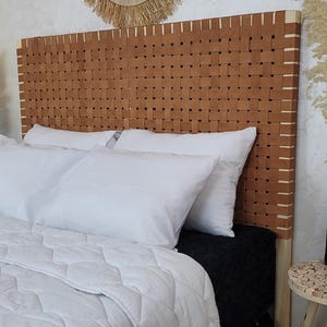 Op de afbeelding: Een bruin geweven leren hoofdbord, gecombineerd met witte kussens en een gewatteerde witte quilt. Een zwarte deken zorgt voor contrast. Het bed staat tegen een neutrale muur. Een ronde spiegel hangt erboven.