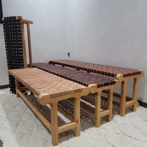May include: Wooden benches with woven leather seats in shades of brown. The benches have light-coloured wooden frames. A tall, black woven panel stands to the left. The benches are arranged in a row.