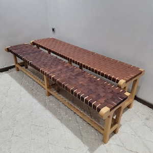Può includere: Due panche in legno con sedute in pelle intrecciata. Le panche hanno una forma rettangolare con gambe in legno marrone chiaro e una seduta in pelle intrecciata marrone scuro. La seconda panca ha una seduta in pelle intrecciata marrone più chiaro.