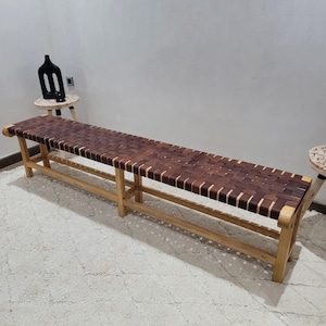 Puede incluir: Un banco largo de madera con un asiento de cuero tejido. El banco tiene un marco de madera marrón claro y un asiento de cuero marrón oscuro. El banco mide aproximadamente 152 cm de largo.
