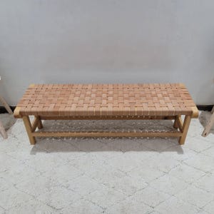 May include: A light brown wooden bench with a woven seat. The bench has a rectangular seat made of woven strips of light brown material. The legs are made of light brown wood and are angled for support. The bench is approximately 122 cm long.
