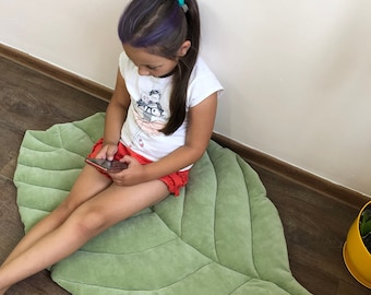 May include: A light green, leaf-shaped floor mat. The mat has a soft, velvety texture and detailed vein patterns. The mat is approximately 40 inches long and 30 inches wide. A child is sitting on the mat.