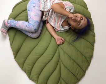 May include: A large, olive green leaf-shaped floor mat with quilted detailing. The mat has a rounded shape and a stem-like extension. The mat is made of a soft, padded material. The mat is designed for children's playrooms or nurseries.