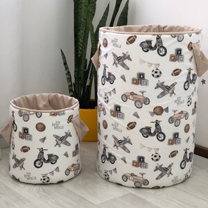 May include: Two fabric storage bins with a cream-colored background and a repeating pattern of vintage vehicles, sports balls, and building blocks. The bins have tan-colored interiors and handles. The text "Retro Boys World" is also printed on the bins.