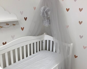 May include: A grey tulle bed canopy featuring a gathered top with a ribbon tie and two matching grey tulle pom-poms hanging from the centre. The sheer fabric drapes downward, creating a soft, layered effect above a white wooden cot.