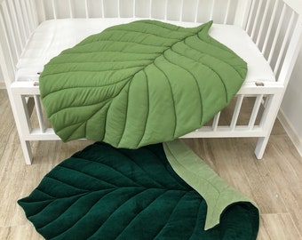 May include: Two leaf-shaped play mats in shades of green. One is a lighter green, resting on a white crib. The other is a darker green, with a lighter green underside. Both mats have quilted detailing.