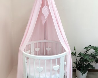 May include: A white oval cot with vertical slats featuring heart-shaped cutouts, topped with a soft pink fabric canopy. The canopy is suspended from a central frame and includes two hanging fabric leaf ornaments in a matching pink colour.