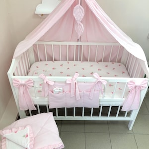 Könnte beinhalten: Ein weißes Kinderbett mit einem rosa Betthimmel, einem rosa Hängeorganizer mit Schleifen und einem Laken mit Blumenmuster. Eine passende rosa Decke mit Rüschen und weißem Blumeneinsatz liegt im Vordergrund und vervollständigt dieses Bettwäscheset.