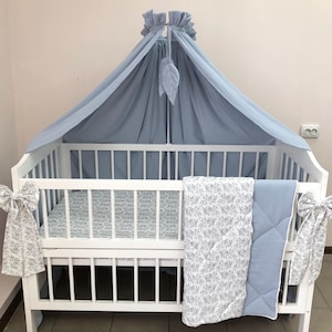 Puede incluir: Una cuna de madera blanca con un dosel de tela azul claro y detalles en forma de hoja. La cuna incluye una funda de colchón y una colcha plegada con un estampado floral en blanco y gris, junto con tela azul claro y lazos decorativos.