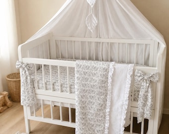 Puede incluir: Una cuna de madera blanca con un dosel blanco transparente y adornos de hojas colgantes. La cuna está vestida con una sábana de estampado floral gris y blanco y una manta a juego con un borde de volantes blancos y dos grandes lazos de tela.
