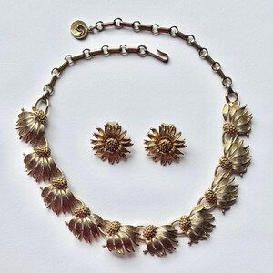May include: A vintage gold-tone jewelry set featuring a necklace and matching clip-on earrings. The necklace and earrings are designed with detailed, stylized flower motifs. The necklace has a chain with a hook clasp. The earrings are in the shape of a flower.
