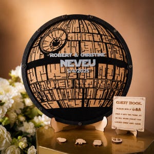 May include: A circular wooden guest book frame shaped like a space station with a black border and natural wood center. It features the text ROBERT & CHRISTINE, NEVEU, and 5/4/2025. A small wooden sign reads GUEST BOOK, PLEASE SIGN A TOKEN, DROP IT INTO THE FRAME.