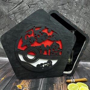 May include: A black wooden box with a hinged lid. The lid features a cutout design of a Pokeball with Pokemon characters. The box is open, revealing a clear glass inside.