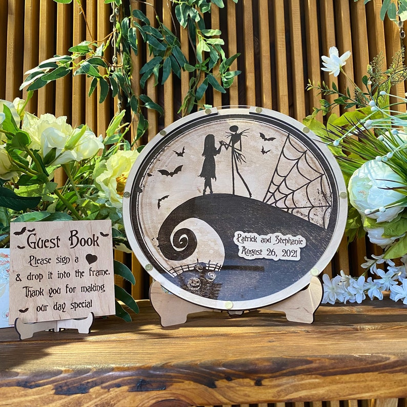 May include: A wooden guest book sign with the text "Guest Book" and instructions to sign and drop it into the frame.  A round wooden sign with a black and white illustration of Jack Skellington and Sally from the Nightmare Before Christmas movie. The sign has the text "Patrick and Stephanie August 26, 2021".