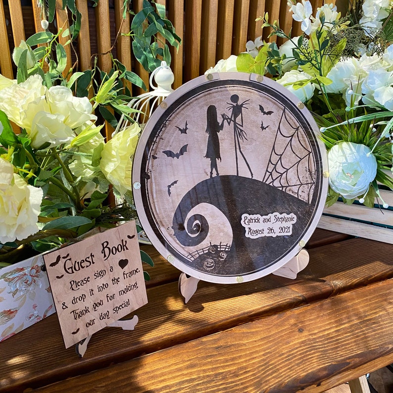 May include: A round wooden guest book with a black and white illustration of Jack Skellington and Sally from the Nightmare Before Christmas. The text "Patrick and Stephanie August 26, 2021" is printed on the guest book.