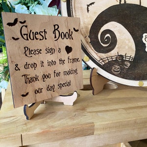 May include: A wooden sign with the text "Guest Book" and instructions to sign a heart and drop it into the frame. The sign is on a wooden stand and has a bat design.