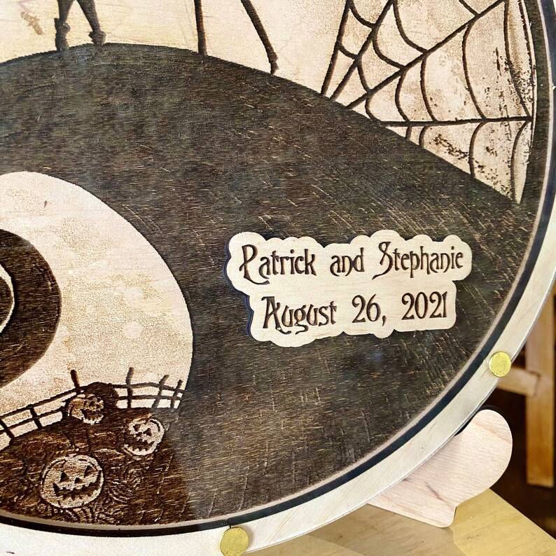 May include: A wooden round sign with a black and white image of a spooky tree and a crescent moon. The sign is engraved with the text "Patrick and Stephanie August 26, 2021".