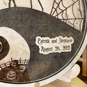 May include: A wooden round plaque with a black and white image of Jack Skellington's head and the words "Patrick and Stephanie August 26, 2021" in a decorative font.