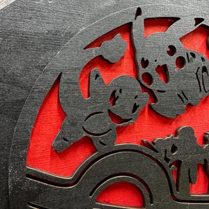 May include: Black wooden wall art featuring a silhouette of three cartoon characters against a red background. The characters are a large, round-eared creature with a wide mouth, a smaller, round-eared creature with a wide mouth, and a smaller, round-eared creature with a wide mouth.