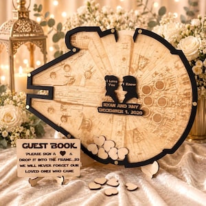 Puede incluir: Un marco de libro de visitas de madera con forma de nave espacial y borde negro. Incluye dos siluetas negras con el texto I Love You e I Know, además de Ryan and Amy December 1, 2020. Un cartel dice GUEST BOOK PLEASE SIGN A HEART & DROP IT INTO THE FRAME.