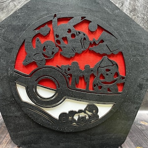 May include: Black wooden wall art featuring a red and white Pokeball with a silhouette of various Pokemon characters, including Bulbasaur, Charmander, Squirtle, and Pikachu.