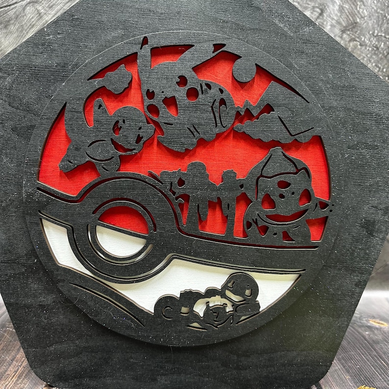 May include: Black wooden wall art featuring a red and white Pokeball with a silhouette of various Pokemon characters, including Bulbasaur, Charmander, Squirtle, and Pikachu.