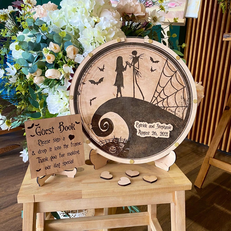 May include: A wooden guest book sign with the text "Guest Book" and instructions to sign and drop into the frame. A wooden round sign with a silhouette of Jack Skellington and Sally from the Nightmare Before Christmas movie. The sign has the text "Patrick and Stephanie August 26, 2021".