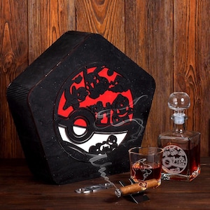 May include: A black wooden box with a red and white Pokeball design. The box is open and a glass decanter with a matching design is visible. A glass with a matching design is also visible. A cigar and a cigar cutter are also visible.