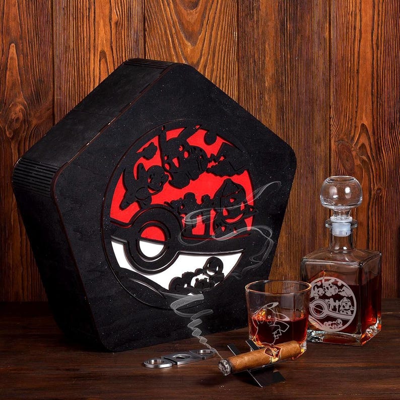 May include: A black wooden box with a red and white Pokeball design. The box is open and a glass decanter with a matching design is visible. A glass with a matching design is also visible. A cigar and a cigar cutter are also visible.