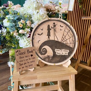 May include: A wooden guest book sign with a black and white silhouette of Jack Skellington and Sally from the Nightmare Before Christmas. The sign reads "Guest Book" and "Please sign a & drop it into the frame. Thank you for making our day special."  The sign is on a wooden stand and there is a round wooden sign with a similar design behind it. The round sign has the text "Patrick and Stephanie August 26, 2021".