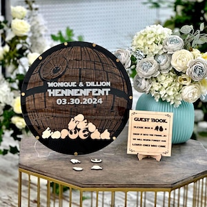 May include: A wooden wedding guest book alternative featuring a Death Star design with the names "MONIQUE & DILLION HENNENFENT" and the date "03.30.2024". Includes a small wooden sign that reads "GUEST BOOK" and a vase of flowers.