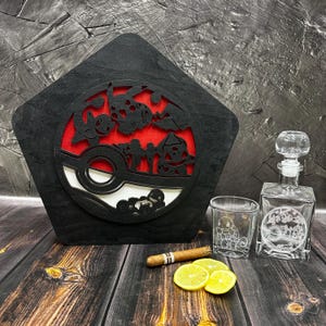 May include: A black wooden box with a red and white Pokeball design. The box is used to hold a glass decanter and two glasses. The decanter and glasses are engraved with a Pokeball design. A cigar and two lemons are also in the image.