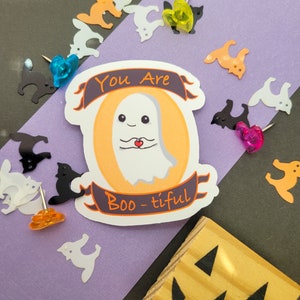 May include: A white ghost with a red heart in its hands, holding a banner that says "You Are Boo-tiful". The ghost is on a yellow background with a black banner.