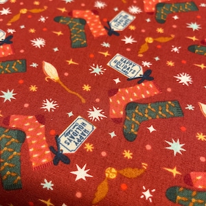 May include: Red fabric with a repeating pattern of Christmas stockings, stars, and wands. The stockings are green and orange with white stripes. The tags on the stockings say "Happy Holidays" and "Season's Greetings".
