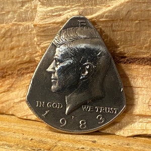 May include: A silver-colored guitar pick shaped like a teardrop with a profile of John F. Kennedy on one side and the words "In God We Trust" and "1983" on the other side.