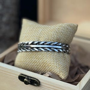 May include: A silver-colored cuff bracelet with a braided design is displayed on a burlap pillow inside a wooden box. The bracelet has a textured pattern and is presented as a jewelry item. The box has a metal clasp.