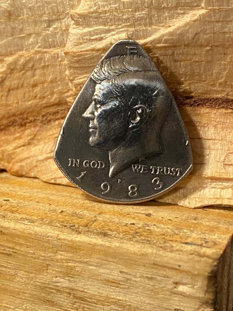 Pu&ograve; includere: Un plettro per chitarra argentato a forma di lacrima con un profilo di John F. Kennedy su un lato e le parole "In God We Trust" e "1983" sull'altro.