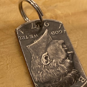 Può includere: Un portachiavi in argento con una moneta da 50 centesimi del 1967 attaccata. La moneta presenta il profilo del viso di un uomo con le parole "In God We Trust" e "Liberty" visibili.