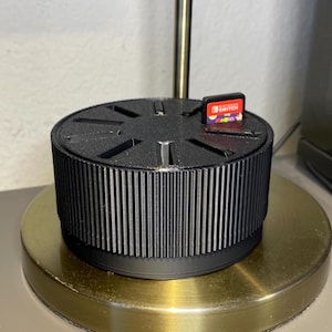 May include: A black plastic game cartridge holder with a gold base. The holder has slots for multiple game cartridges and a raised, textured rim. A Nintendo Switch game cartridge is visible in one of the slots.
