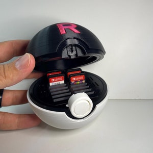 May include: A black and white Poke Ball-shaped case with a red "R" on the top. The case is open and has slots for Nintendo Switch game cartridges. The case is being held in a hand.