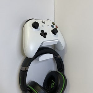 May include: A white Xbox controller with black buttons and a green and black headset hanging from it. The controller and headset are mounted on a white wall mount.