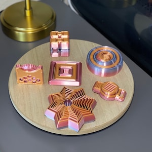 May include: A collection of six small, decorative, metallic objects in shades of copper and rose gold. The objects are arranged on a wooden platform. The objects include a cube, a square, a circle, a star, and two other geometric shapes.