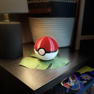 May include: A red and white Poke Ball toy on a green leaf. The Poke Ball is a popular collectible toy from the Pokemon franchise.