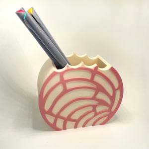 May include: A decorative pen holder with a cream-colored base and a pink, geometric design. The holder contains three gray pens with colorful tips. The holder has a unique, rounded shape and a scalloped top edge.
