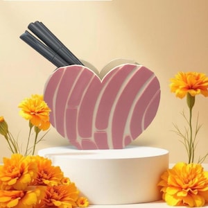 May include: A pink heart-shaped desk organizer with white accents, holding two dark gray pens. The organizer sits on a white pedestal, surrounded by vibrant orange marigold flowers. The background is a soft, warm beige.