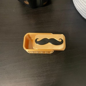 May include: A small, gold-colored box with a hinged lid. The lid has a black mustache design.