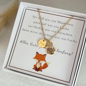 Könnte beinhalten: Goldfarbene Halskette mit einem Fuchs-Anhänger und einem runden Charm mit dem Namen "Paul" und einem Herz. Die Halskette wird in einer weißen Box mit einer Cartoon-Fuchs-Illustration und deutschem Text präsentiert.