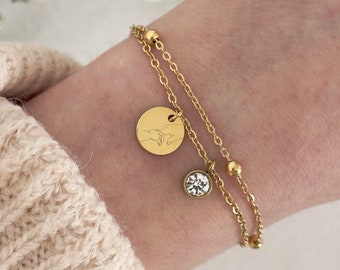 May include: A gold-colored double-strand bracelet with a round charm and a small, clear crystal. The charm has an engraved design. The bracelet is worn on a wrist, with a beige sweater in the background.
