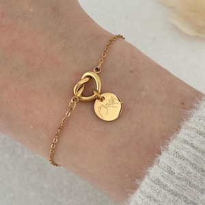 Könnte beinhalten: Ein goldenes Kettenarmband mit einem Knotenanhänger und einem runden Anhänger, der mit einem Bild von zwei Händen, die ein Pinky-Versprechen machen, graviert ist.